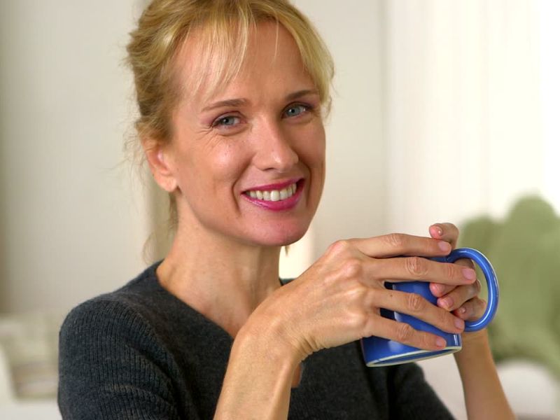 Žena plave kose sa rozim karminom na usnama i u tamnoj majici nasmejano drži šolju čaja u rukama
